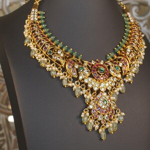 May include: A gold-toned necklace featuring intricate metalwork, embedded with red, green, and clear stones. The design includes multiple rows of small green beads along the top edge and dangling pale green teardrop beads along the bottom.