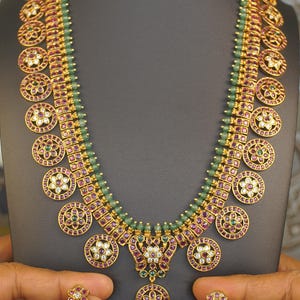 May include: A gold-toned necklace and earring set featuring circular motifs inlaid with red and green stones and clear crystals. The necklace has a strand of small green beads, while the earrings include dangling green teardrop-shaped beads.