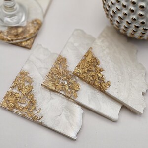 May include: Four white square coasters with gold leaf accents. The coasters have a slightly irregular shape and a white background with gold leaf flakes scattered across the surface.
