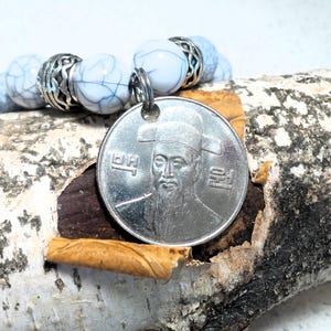 May include: A white and blue marble bracelet with a silver charm featuring a Korean Won coin with a portrait of King Sejong the Great. The coin has the Korean text "대한민국" on it.