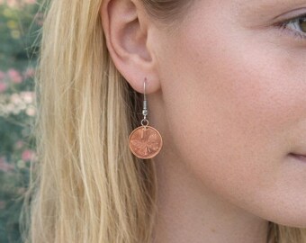 May include: A copper-colored coin earring with a butterfly design is suspended from a silver-colored hook. The earring is round and has a detailed image of a butterfly. The background is blurred, with a person's hair and face visible.
