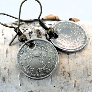 May include: A pair of silver coin earrings with a bronze hook. The coins are from 1956 and feature the text "ONE PIE" and a stylized image of a bird.