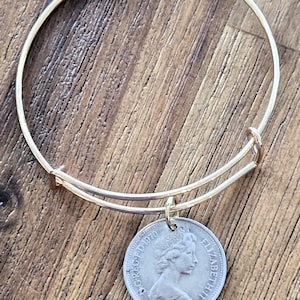 Puede incluir: Un brazalete dorado con un colgante de moneda plateada. La moneda presenta un perfil de mujer y el texto "ELIZABETH II" y "1970". El brazalete está sobre una superficie de madera.