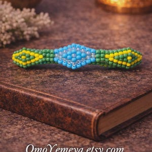 Può includere: Un fermaglio per capelli con perline nei toni del blu, verde e giallo, appoggiato su un libro rilegato in pelle marrone. Il fermaglio presenta una forma a diamante blu centrale, affiancata da sezioni verdi e gialle. Il nome del negozio Etsy, OmoYemeya.etsy.com, è visibile.