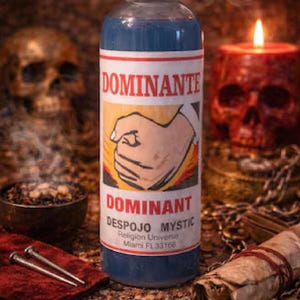 May include: A blue bottle with a white and red label that reads "DOMINANTE" and "DESPOJO MYSTIC". The label features a graphic of a clenched fist. The background includes a skull-shaped candle and a human skull.