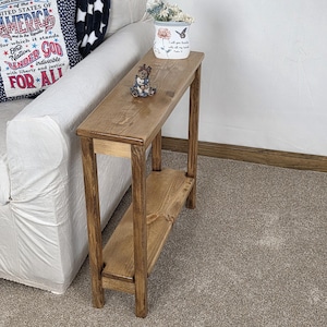 May include: A narrow, rectangular wooden console table with a medium brown stain, featuring a flat top surface and a lower shelf supported by four straight legs. The table is constructed from wood with visible grain patterns throughout.