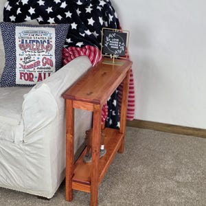 May include: A wooden console table with a lower shelf, stained in a warm reddish-brown hue. A decorative pillow with patriotic text and a blanket with white stars on a navy background are visible. A small sign reads "LOVE MAKES A HOUSE A HOME."