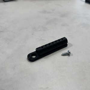 May include: A black plastic cable organiser with a screw. The organiser has multiple holes for cables and wires.