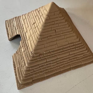 May include: A tan-coloured, miniature pyramid with a textured brick pattern. The pyramid has multiple stepped levels and a pointed top. The structure includes a small, arched opening on one side, suggesting a doorway or entrance.