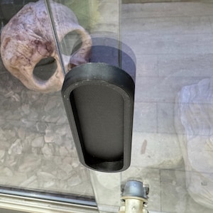 May include: A black, rectangular aquarium accessory with a textured interior surface. The accessory is mounted on a glass panel, with a blurred background featuring decorative rocks and a light-colored structure.
