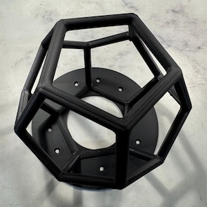 May include: A black geometric sculpture featuring a dodecahedron cage design atop a circular base with multiple holes. The sculpture is made of a matte black material and is set against a light grey surface.