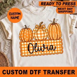 May include: A cream-colored t-shirt featuring a graphic of three orange pumpkins above a scalloped rectangular banner with an orange and white gingham pattern. The name Olivia is printed in black cursive script across the center of the banner.
