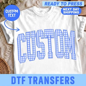 May include: A white short-sleeved t-shirt featuring the word CUSTOM printed in a large, arched, blue and white gingham check pattern across the chest. The shirt has a crew neckline and a standard fit, showcasing a clean, simple design.