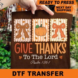 May include: A dark brown t-shirt featuring a graphic with three rectangular panels containing a brown bow, an orange pumpkin, and an orange bow, all on a striped background. The text reads GIVE THANKS To The Lord Psalm 136:1 with autumn leaves.