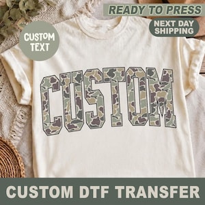 May include: A cream-colored short-sleeve t-shirt featuring the word CUSTOM in large, block-style letters across the chest. The letters are filled with a multi-colored camouflage pattern in shades of green, brown, tan, and black.
