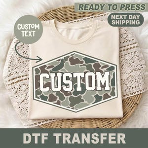May include: A cream-colored t-shirt featuring a central hexagonal graphic with a green, brown, and tan camouflage pattern. The word CUSTOM is printed in large, bold white block letters across the center of the camouflage hexagon design.