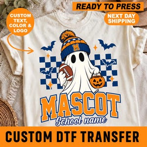 May include: A cream-colored t-shirt featuring a graphic of a ghost wearing a blue and orange beanie with a letter M, holding a football and a jack-o'-lantern bucket. The design includes a blue and white checkered background, bats, and the text MASCOT School name.