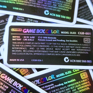 May include: Close-up of a black Game Boy Color Model OLED sticker with holographic text and a checkmark. The sticker includes the text "GAME BOY.COLOR MODEL OLED. CGB-001", "RATING: DC3V 0.6W", "BATTERY: LR6 (AA)x2", "1998 Nintendo", "Patents issued and Pending. See booklet.", "THIS DEVICE COMPLIES WITH PART 15 OF THE FCC RULES. OPERATION IS SUBJECT TO THE FOLLOWING TWO CONDITIONS: (1) THIS DEVICE MAY NOT CAUSE HARMFUL INTERFERENCE, AND (2) THIS DEVICE MUST ACCEPT ANY INTERFERENCE RECEIVED, INCLUDING INTERFERENCE THAT MAY CAUSE UNDESIRED OPERATION. THIS CLASS B DIGITAL APPARATUS COMPLIES WITH CANADIAN ICES 003", "MADE IN USA", "C/CGB-USA-1", and "ACN 060 566 083".