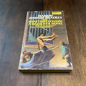 May include: A well-worn paperback copy of "The Shattered Chain: A Darkover Novel" by Marion Zimmer Bradley. The cover features a fantasy illustration of sword-wielding figures. The book's spine is yellowed, indicating its age.