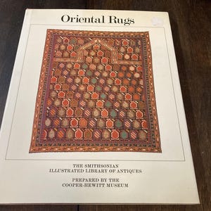 Op de afbeelding: Een boek getiteld "Oriental Rugs" met een gedetailleerde afbeelding van een tapijt op de omslag. Het tapijtontwerp heeft een herhalend patroon van rode, oranje en groene vormen op een donkere achtergrond. Het boek maakt deel uit van de Smithsonian Illustrated Library of Antiques.