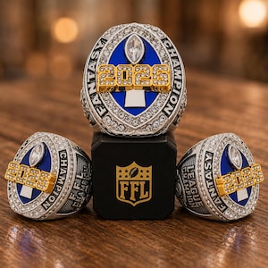 May include: Three silver-toned championship rings featuring a blue oval center with a clear stone, gold-colored 2025 text, and clear crystals along the border. The rings are displayed with a black square base marked with a gold FFL logo.