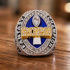 May include: A silver-toned championship ring featuring a blue oval centre with a central football icon and the gold-coloured text 2025 encrusted with clear stones. The border is lined with small clear crystals and the words FANTASY CHAMPION.