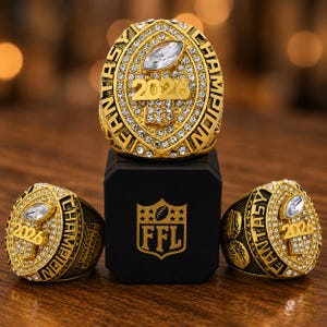 May include: Three gold-toned fantasy football championship rings featuring clear rhinestones, a central football-shaped gem, and the text 2026 FANTASY CHAMPION. The rings are displayed with a black box featuring a gold FFL shield logo and football.