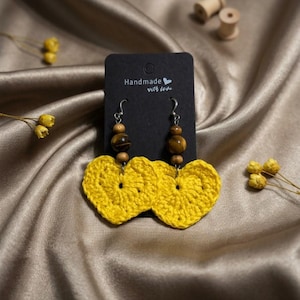 May include: A pair of dangle earrings featuring bright yellow crocheted hearts suspended from silver-toned hooks. Each earring includes a brown wooden bead and a tiger's eye gemstone bead. The earrings are attached to a black card labeled Handmade with love.