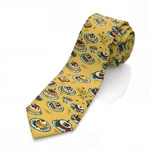 May include: A mustard yellow necktie featuring a repeating pattern of various cakes, tarts, and small teacups on plates. The illustrations include round cakes with berries, square pastries, and small floral sprigs scattered across the fabric surface.