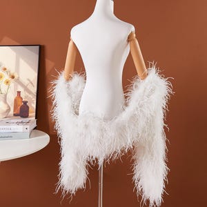 May include: A white feather boa draped over a mannequin. The boa is made of long, fluffy feathers. The mannequin is white with wooden arms and is on a metal stand. The background is a solid brown color.