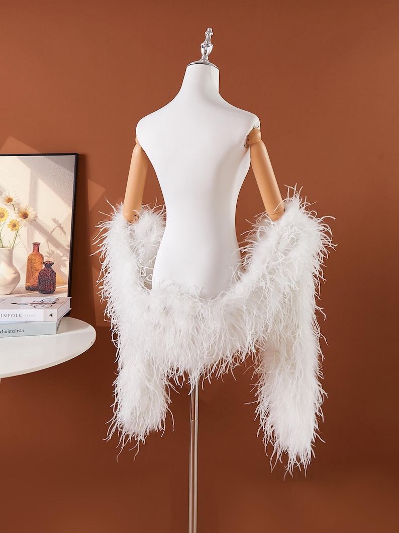 May include: A white feather boa draped over a mannequin. The boa is made of long, fluffy feathers. The mannequin is white with wooden arms and is on a metal stand. The background is a solid brown color.