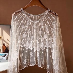 May include: A sheer, off-white lace capelet featuring intricate floral embroidery and delicate beadwork. The garment has a scalloped hemline and a V-neckline, with vertical beaded lines radiating from the neckline down to the edges of the fabric.