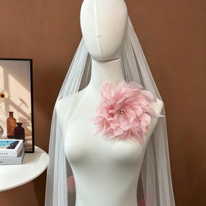 K&ouml;nnte beinhalten: Ein wei&szlig;er Brautschleier, der &uuml;ber eine Schaufensterpuppe drapiert ist und eine gro&szlig;e, zartrosa Federblume auf der Brust aufweist. Die Blume ist mit kleinen, hellen Perlen verziert und der Schleier besteht aus transparentem, leichtem T&uuml;llstoff.