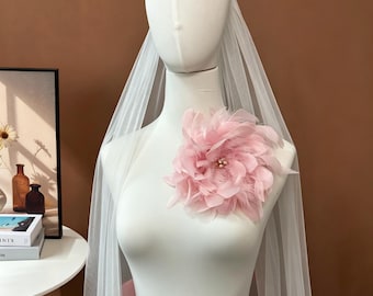 May include: A white bridal veil draped over a mannequin, featuring a large, soft pink feather flower embellishment at the chest. The flower is centered with small, light-colored beads, and the veil consists of sheer, lightweight tulle fabric.