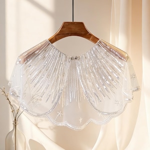 May include: A white, sheer capelet with silver sequins and beaded fringe, hanging on a wooden hanger. The capelet has a scalloped edge and is decorated with star-shaped embellishments. The background includes a vase with a plant and a draped fabric.