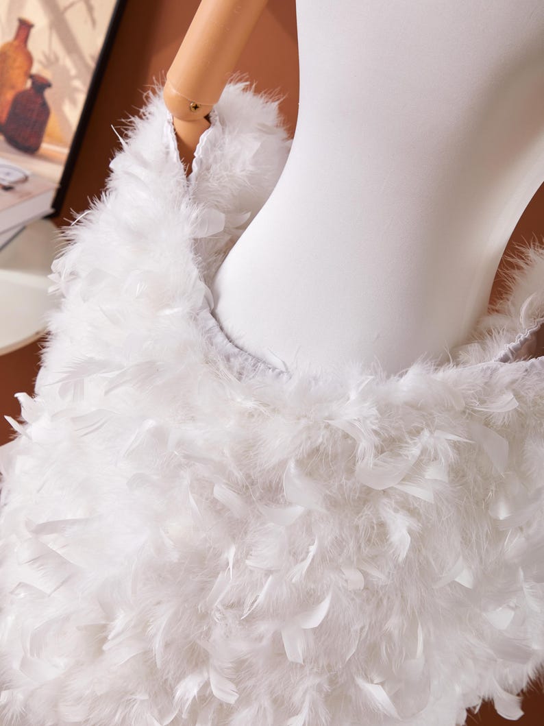 May include: A white, feathery garment, possibly a shawl or wrap, is draped over a mannequin. The feathers are densely packed, creating a fluffy texture. The garment appears to be part of a costume or fashion piece.