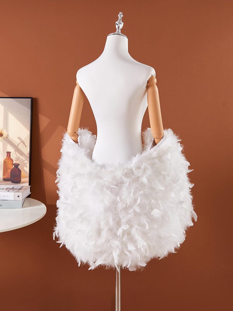 May include: A white, fluffy feather skirt is displayed on a mannequin. The skirt is made of soft, white feathers and is designed to be worn around the waist. The mannequin has wooden arms and is set against a brown background.