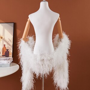 May include: A white feather boa draped over a mannequin. The boa is made of long, soft feathers and is displayed against a brown background. The mannequin has wooden arms and a white torso.