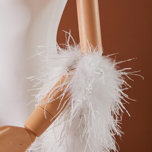 May include: A white, fluffy feather boa or cuff detail, possibly for a garment, is wrapped around a mannequin arm. The feathers are long and soft, creating a voluminous texture. The background is a warm, neutral brown.