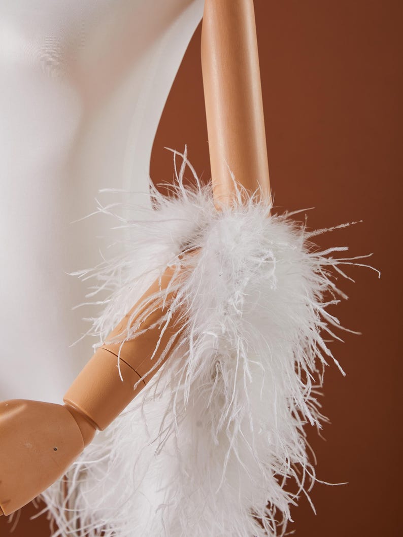 May include: A white, fluffy feather boa or cuff detail, possibly for a garment, is wrapped around a mannequin arm. The feathers are long and soft, creating a voluminous texture. The background is a warm, neutral brown.