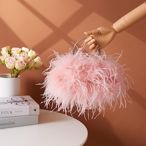 May include: A pink feather handbag with a silver handle is held by a mannequin hand. The bag has a fluffy texture. A bouquet of roses and books are on a white table. The background is a warm, neutral tone.