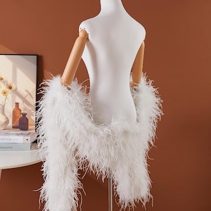 May include: A white feather boa draped over a mannequin. The boa is made of long, soft, fluffy feathers. The mannequin is white with wooden arms. The background is a solid brown color.