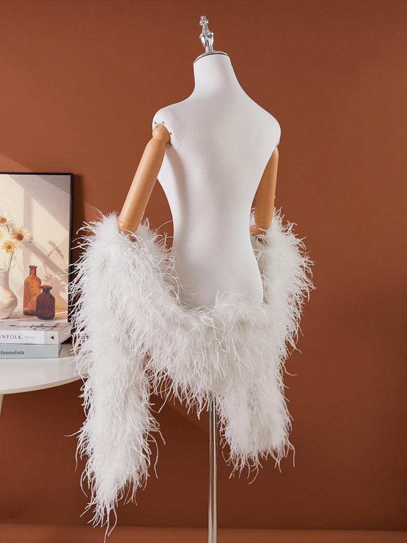 May include: A white feather boa draped over a mannequin. The boa is made of long, soft, fluffy feathers. The mannequin is white with wooden arms. The background is a solid brown color.