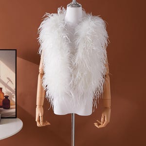 Könnte beinhalten: Eine weiße Federweste auf einer Schaufensterpuppe. Die Weste besteht aus langen, weichen weißen Federn, die ihr eine flauschige Textur verleihen. Die Schaufensterpuppe hat Holzärmel und steht vor einem braunen Hintergrund.