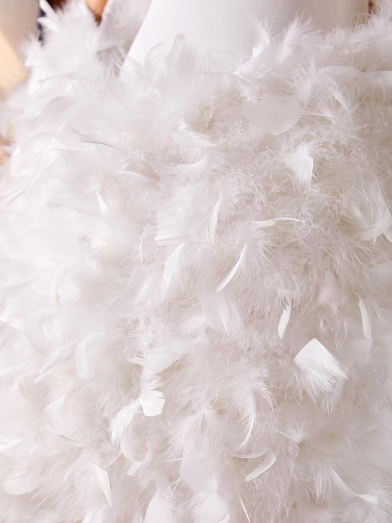 May include: Close-up of a white garment adorned with numerous fluffy feathers. The feathers are layered, creating a soft, textured appearance. The overall effect is light and airy, suggesting a formal or decorative piece.