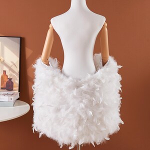 May include: A white feather skirt displayed on a mannequin. The skirt is made of soft, fluffy white feathers, creating a voluminous and textured appearance. The mannequin has wooden arms and a white torso, set against a warm brown background.