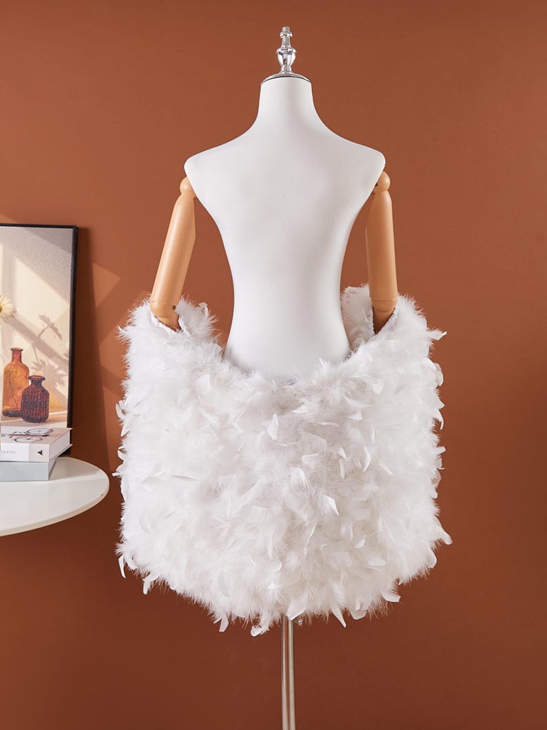 May include: A white feather skirt displayed on a mannequin. The skirt is made of soft, fluffy white feathers, creating a voluminous and textured appearance. The mannequin has wooden arms and a white torso, set against a warm brown background.