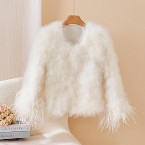 May include: A cream-coloured, fluffy jacket with a rounded neckline hangs from a wooden hanger. The jacket has a soft, textured appearance, with long, feathery accents at the cuffs. The garment is displayed against a neutral background, suggesting a focus on the clothing item.