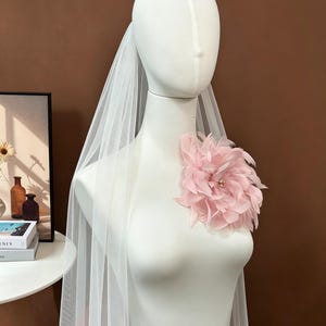 K&ouml;nnte beinhalten: Ein wei&szlig;er T&uuml;llschleier, der &uuml;ber eine Schaufensterpuppe drapiert ist und auf der linken Seite mit einer gro&szlig;en, geschichteten blassrosa Federblume verziert ist. Die Blume hat in der Mitte eine kleine Ansammlung winziger, heller Perlen.