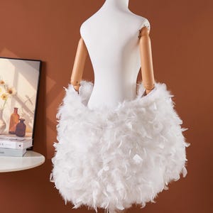 May include: A white dress form with wooden arms is adorned with a skirt made of fluffy white feathers. The dress form is on a silver stand, set against a warm brown background. A framed picture and books are visible in the background.